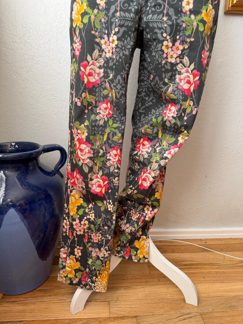Puede incluir: Un pantal&oacute;n gris oscuro con un vibrante estampado floral de rosas rojas, rosas y amarillas, hojas verdes y un sutil fondo de damasco negro. El pantal&oacute;n se muestra sobre un soporte blanco, exhibiendo el dise&ntilde;o de pierna larga.