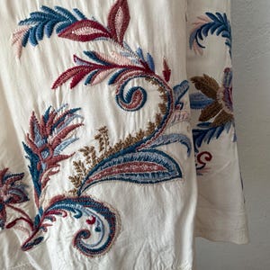 May include: Close-up of a cream-colored fabric with intricate embroidery. The embroidery features a paisley design with blue, red, and brown floral patterns. The fabric has a textured appearance and a scalloped edge.