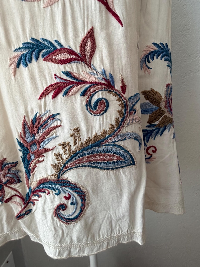 May include: Close-up of a cream-colored fabric with intricate embroidery. The embroidery features a paisley design with blue, red, and brown floral patterns. The fabric has a textured appearance and a scalloped edge.