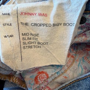 Op de afbeelding: Close-up van een stoffen label van een blauwe jeans. Het label toont de merknaam "JOHNNY WAS" in het rood. De stijl is "THE CROPPED BABY BOOT". Andere details zijn "MID RISE", "SLIM FIT", "SLIGHT BOOT" en "STRETCH".