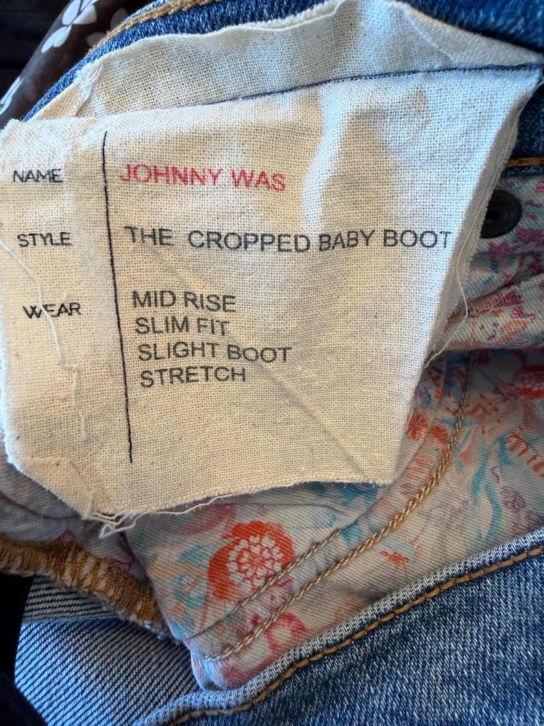 Op de afbeelding: Close-up van een stoffen label van een blauwe jeans. Het label toont de merknaam "JOHNNY WAS" in het rood. De stijl is "THE CROPPED BABY BOOT". Andere details zijn "MID RISE", "SLIM FIT", "SLIGHT BOOT" en "STRETCH".