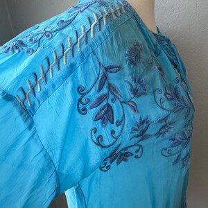 Puede incluir: Una blusa azul claro con intrincados bordados en tonos azules y blancos. El dise&ntilde;o floral y de hojas se presenta en las mangas y el corpi&ntilde;o. La blusa tiene un cuello redondo y mangas largas.