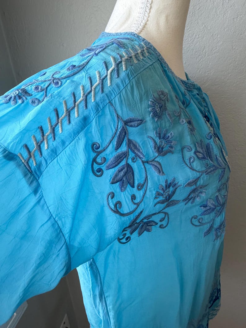 Puede incluir: Una blusa azul claro con intrincados bordados en tonos azules y blancos. El dise&ntilde;o floral y de hojas se presenta en las mangas y el corpi&ntilde;o. La blusa tiene un cuello redondo y mangas largas.