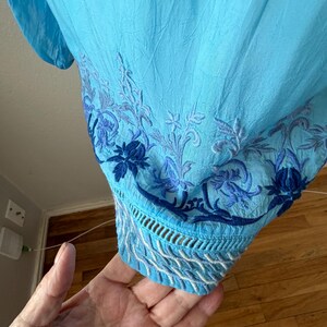 Puede incluir: Una blusa azul claro con intrincados bordados florales en tonos azules. El dobladillo inferior presenta un dise&ntilde;o calado detallado con costuras blancas y azules. La tela parece ligera y texturizada.
