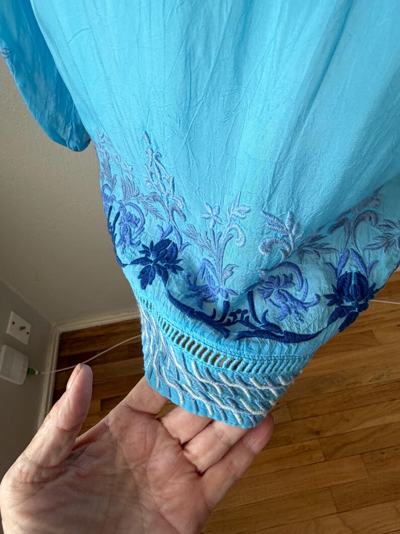 Puede incluir: Una blusa azul claro con intrincados bordados florales en tonos azules. El dobladillo inferior presenta un dise&ntilde;o calado detallado con costuras blancas y azules. La tela parece ligera y texturizada.