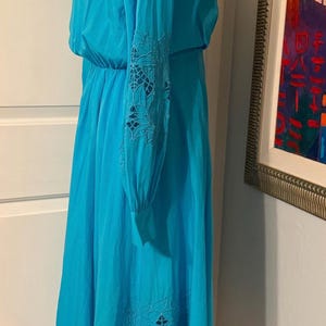 Puede incluir: Un vestido turquesa vibrante con mangas largas y una falda fluida. Las mangas y la falda presentan intrincados detalles florales calados. El vestido tiene cintura el&aacute;stica y un escote modesto, perfecto para el verano.