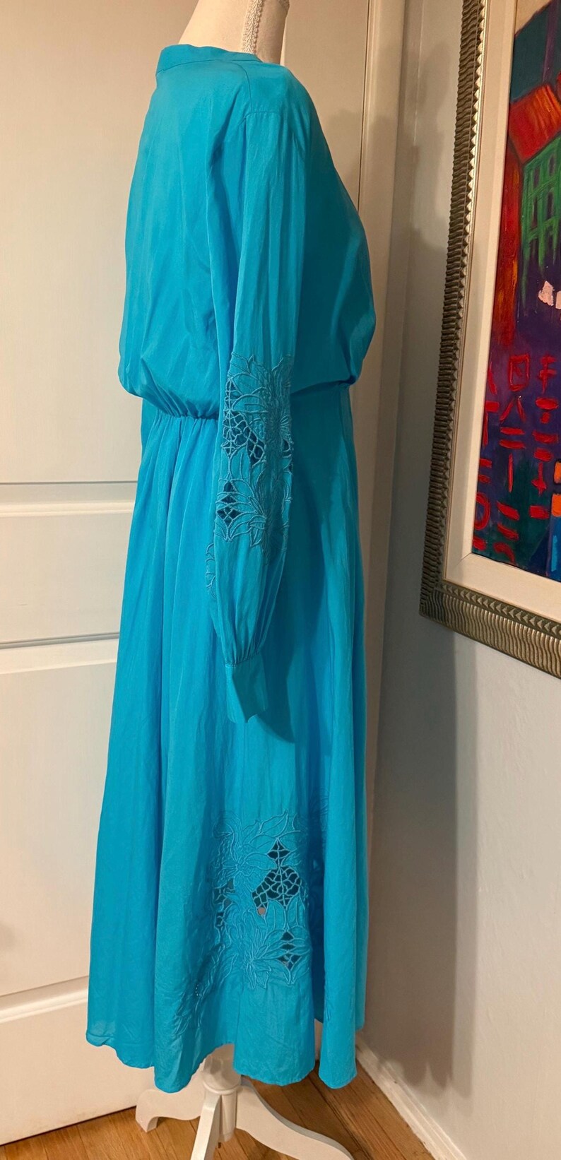 Puede incluir: Un vestido turquesa vibrante con mangas largas y una falda fluida. Las mangas y la falda presentan intrincados detalles florales calados. El vestido tiene cintura el&aacute;stica y un escote modesto, perfecto para el verano.