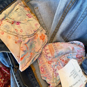Op de afbeelding: Close-up van een blauwe spijkerbroek met bloemenpatroon voering van de zak. De jeans heeft een ritssluiting en een knoopsluiting. Een wit label met tekst is zichtbaar. De jeans is gelabeld "The Cropped Baby Boot".