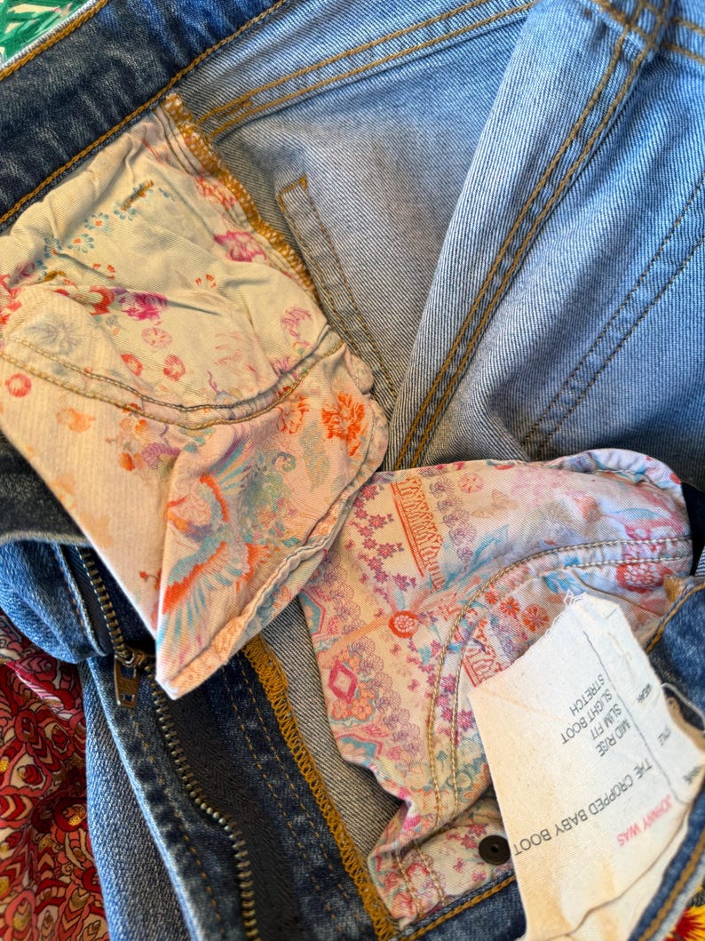 Op de afbeelding: Close-up van een blauwe spijkerbroek met bloemenpatroon voering van de zak. De jeans heeft een ritssluiting en een knoopsluiting. Een wit label met tekst is zichtbaar. De jeans is gelabeld "The Cropped Baby Boot".