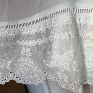 May include: Close-up of white fabric with intricate embroidered details. The design includes a scalloped edge, eyelet cutouts, and floral and butterfly motifs. The fabric appears to be lightweight and slightly textured.