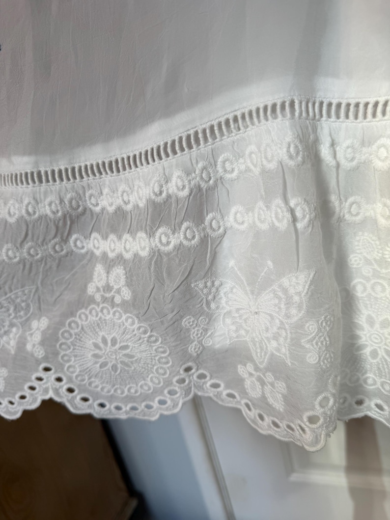 May include: Close-up of white fabric with intricate embroidered details. The design includes a scalloped edge, eyelet cutouts, and floral and butterfly motifs. The fabric appears to be lightweight and slightly textured.