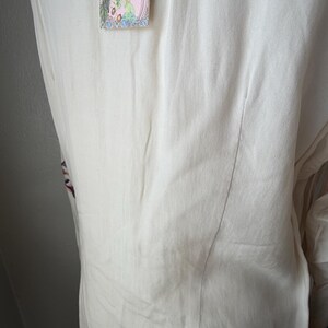 May include: Off-white, flowing top with floral embroidery and a decorative tag. The tag features a pink background with colorful floral and botanical illustrations. The top has a vertical seam down the back.