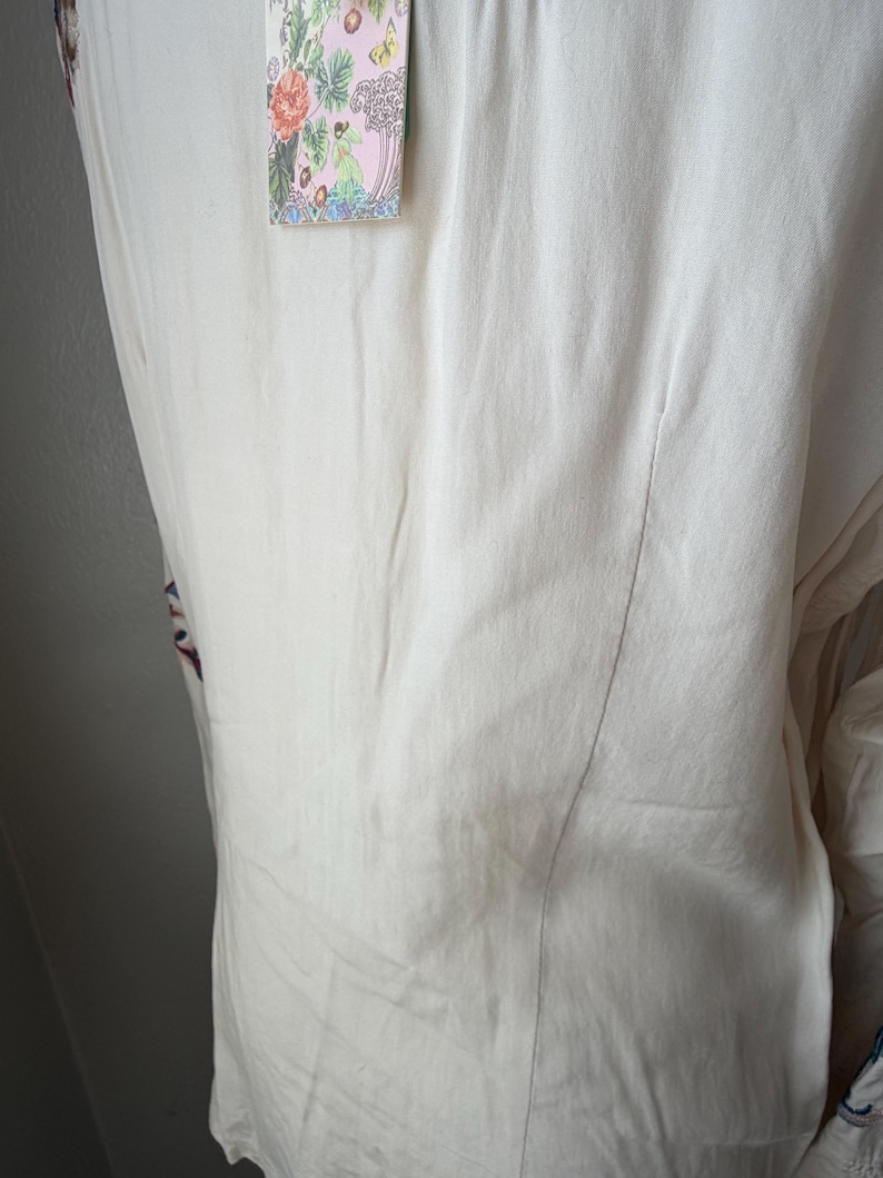 May include: Off-white, flowing top with floral embroidery and a decorative tag. The tag features a pink background with colorful floral and botanical illustrations. The top has a vertical seam down the back.