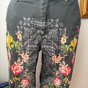 Puede incluir: Pantalones de color gris oscuro con un llamativo patr&oacute;n de damasco negro sobre la tela. El dise&ntilde;o est&aacute; acentuado con ilustraciones florales vibrantes en tonos rosa, amarillo y verde, que se extienden desde el dobladillo hasta la cintura.