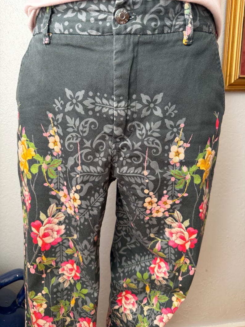 Puede incluir: Pantalones de color gris oscuro con un llamativo patr&oacute;n de damasco negro sobre la tela. El dise&ntilde;o est&aacute; acentuado con ilustraciones florales vibrantes en tonos rosa, amarillo y verde, que se extienden desde el dobladillo hasta la cintura.