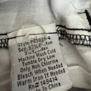 Puede incluir: Una etiqueta de cuidado de prenda blanca con un bot&oacute;n de pl&aacute;stico transparente y el texto impreso: Style: P63821-4, 97% Cotton, 3% Spandex, Machine Wash Cold, Tumble Dry Low, Only Non Chlorine Bleach When Needed, Warm Iron If Needed, Rn# 149414, Made In China.
