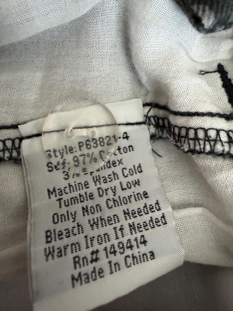 Puede incluir: Una etiqueta de cuidado de prenda blanca con un bot&oacute;n de pl&aacute;stico transparente y el texto impreso: Style: P63821-4, 97% Cotton, 3% Spandex, Machine Wash Cold, Tumble Dry Low, Only Non Chlorine Bleach When Needed, Warm Iron If Needed, Rn# 149414, Made In China.