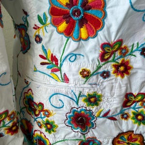 Puede incluir: Chaqueta blanca con bordados florales coloridos. Los bordados presentan flores y hojas rojas, amarillas, azules y verdes. La chaqueta tiene un bolsillo y est&aacute; hecha de tela ligera.