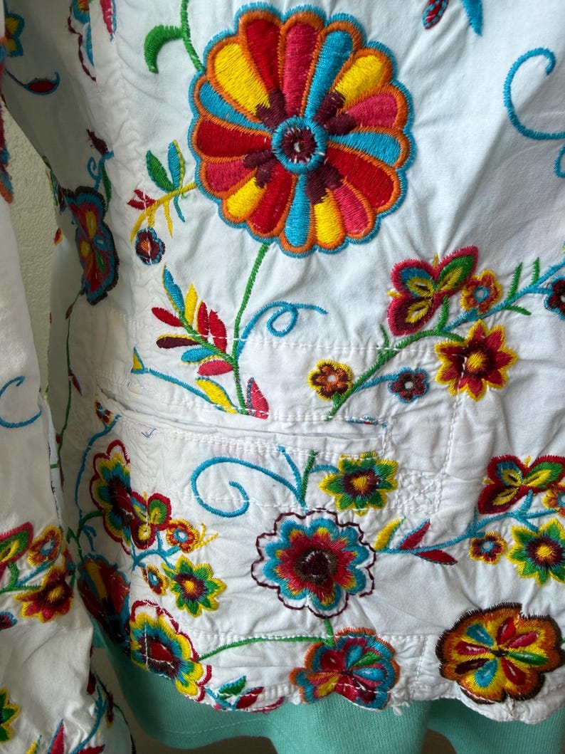 Puede incluir: Chaqueta blanca con bordados florales coloridos. Los bordados presentan flores y hojas rojas, amarillas, azules y verdes. La chaqueta tiene un bolsillo y est&aacute; hecha de tela ligera.