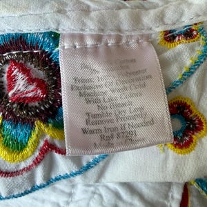 Puede incluir: Primer plano de una tela blanca con bordados florales coloridos en rojo, azul, amarillo y verde. Una etiqueta rosa con texto est&aacute; adherida a la tela. El bordado es detallado y vibrante, con una variedad de puntadas.