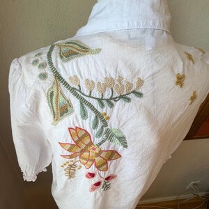 May include: White button-down shirt with colorful floral embroidery on the back. The embroidery features a large flower with red, yellow, and orange petals, along with green leaves and stems. The shirt is displayed on a mannequin.