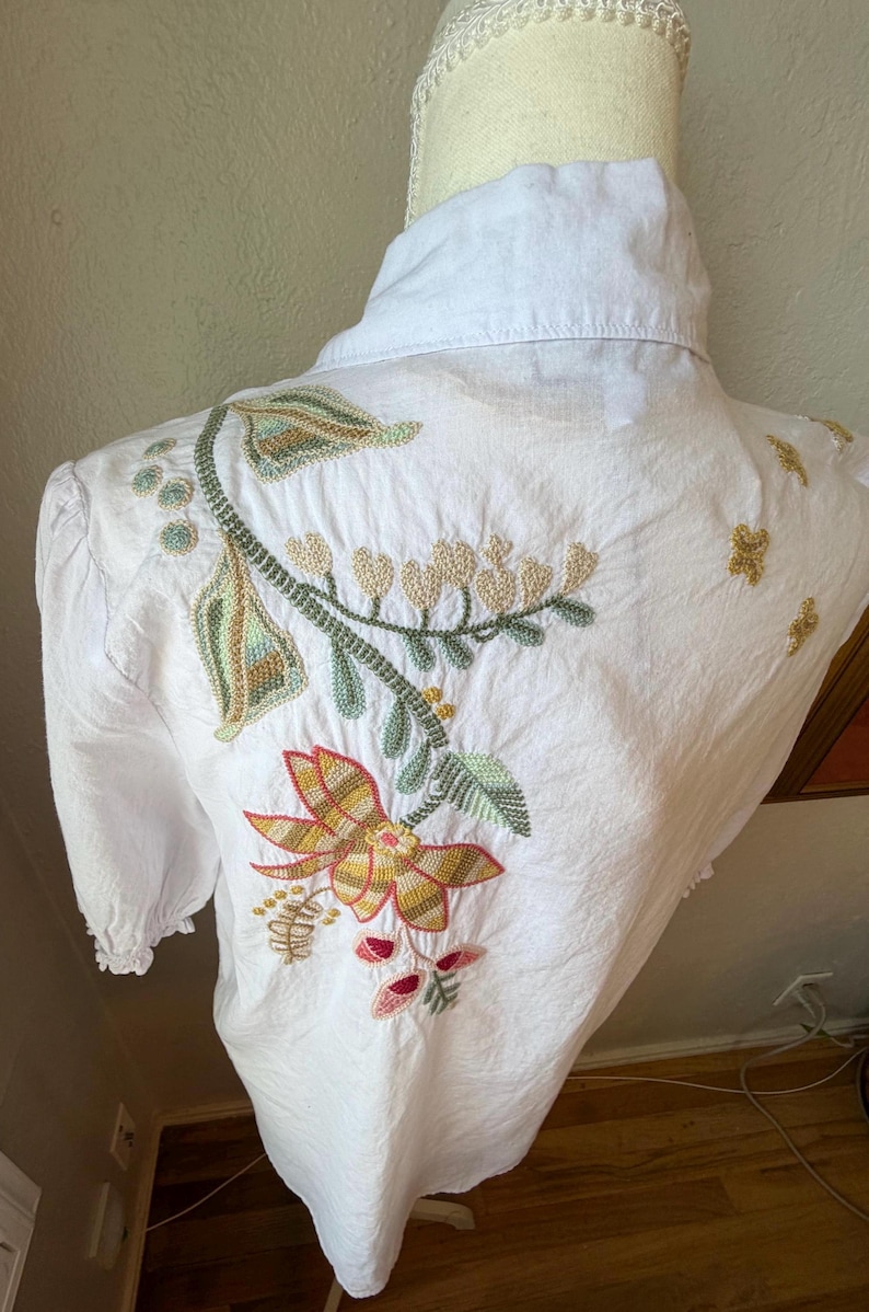 May include: White button-down shirt with colorful floral embroidery on the back. The embroidery features a large flower with red, yellow, and orange petals, along with green leaves and stems. The shirt is displayed on a mannequin.