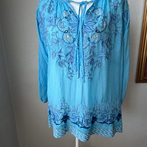 Puede incluir: Una t&uacute;nica azul claro con mangas largas y bordados florales detallados en tonos azules y morados. La parte superior presenta un dise&ntilde;o de cuello atado y bordados en el escote y el dobladillo.