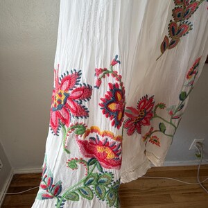 May include: White dress with colorful floral embroidery. The embroidery features red, pink, yellow, and blue flowers with green leaves. The dress has vertical pleats and a scalloped hem.