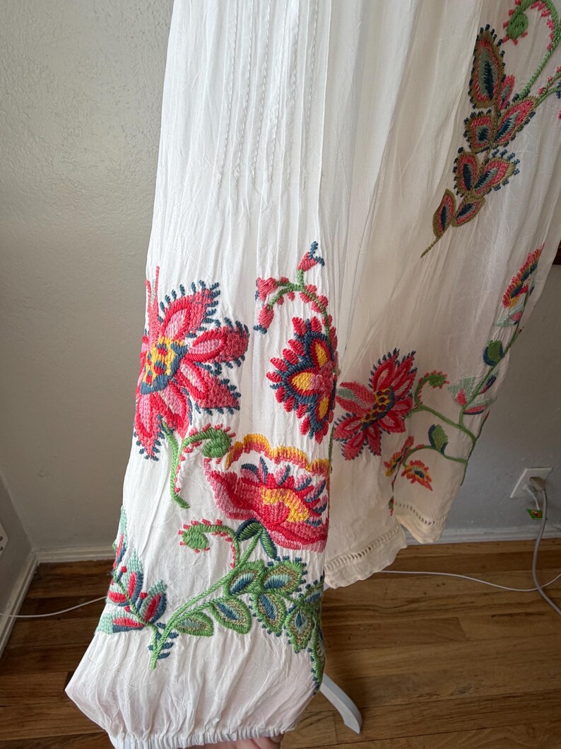 May include: White dress with colorful floral embroidery. The embroidery features red, pink, yellow, and blue flowers with green leaves. The dress has vertical pleats and a scalloped hem.