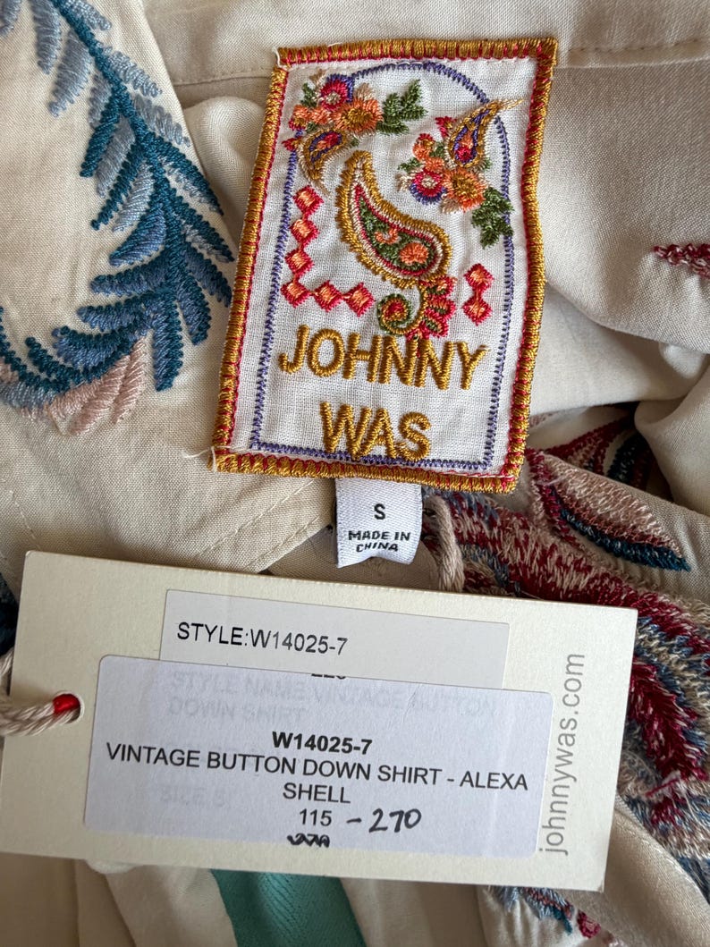 May include: Cream-colored vintage button-down shirt with colorful embroidered details. The shirt features a tag with the brand name "JOHNNY WAS" and the style information. The shirt is size small.
