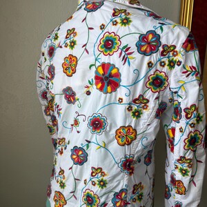 Puede incluir: Camisa blanca con bordado floral colorido. El bordado presenta flores rojas, amarillas, azules y verdes con vides verdes. La camisa tiene mangas largas y cuello.