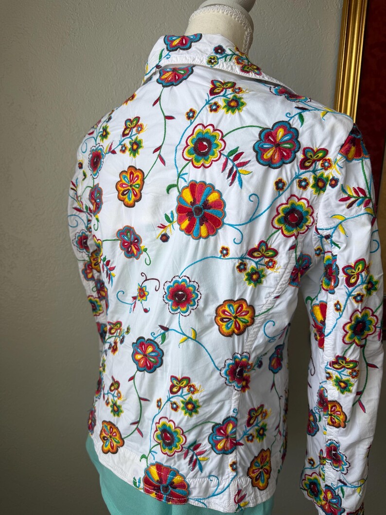 Puede incluir: Camisa blanca con bordado floral colorido. El bordado presenta flores rojas, amarillas, azules y verdes con vides verdes. La camisa tiene mangas largas y cuello.