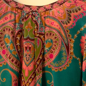 May include: Close-up of a teal-colored top with a vibrant paisley pattern in pink, green, and brown. The neckline is trimmed with a floral design. The fabric appears soft and flowing.