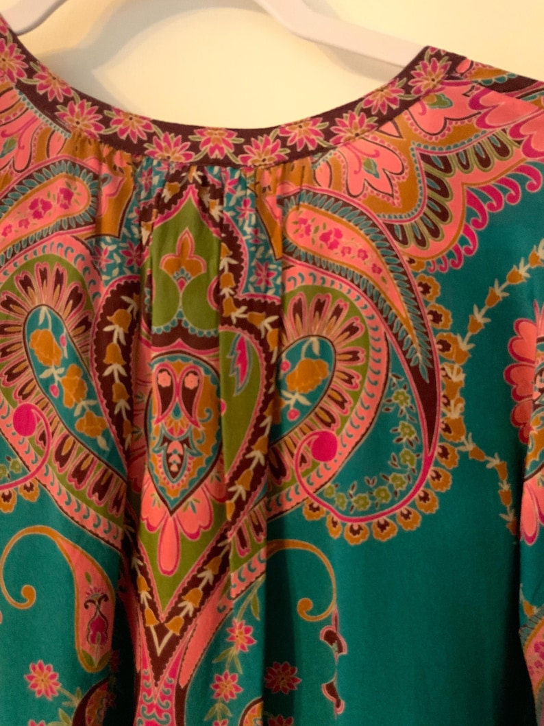 May include: Close-up of a teal-colored top with a vibrant paisley pattern in pink, green, and brown. The neckline is trimmed with a floral design. The fabric appears soft and flowing.