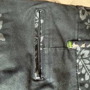Puede incluir: Un pantal&oacute;n negro con un bolsillo de ojal vertical y una cinturilla adornada con un estampado floral gris oscuro. La tela muestra un estampado bot&aacute;nico repetitivo de hojas y flores, con un peque&ntilde;o toque de verde y rosa visible.