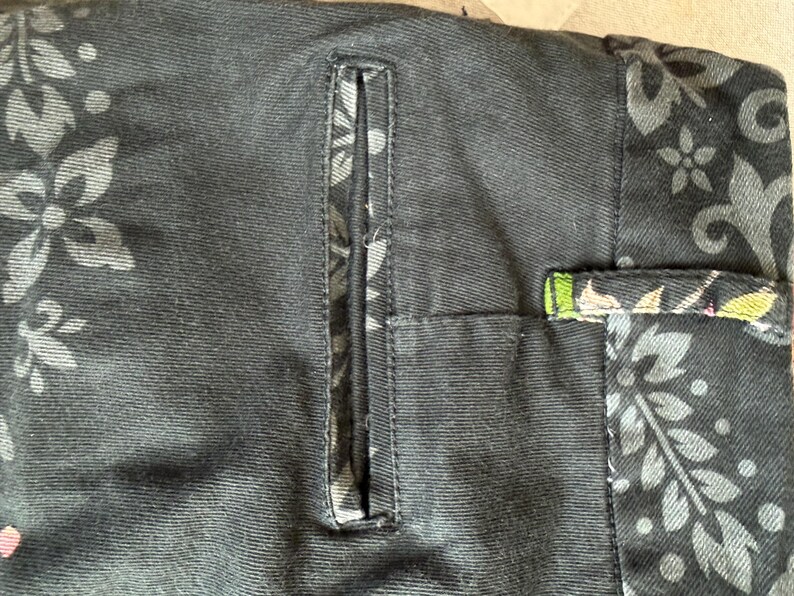 Puede incluir: Un pantal&oacute;n negro con un bolsillo de ojal vertical y una cinturilla adornada con un estampado floral gris oscuro. La tela muestra un estampado bot&aacute;nico repetitivo de hojas y flores, con un peque&ntilde;o toque de verde y rosa visible.