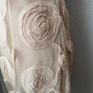 May include: Close-up of a cream-colored fabric with textured, swirling floral appliqu&eacute;s. The fabric has a crinkled appearance, and the appliqu&eacute;s are made of the same material, creating a cohesive design. The overall aesthetic is soft and delicate.