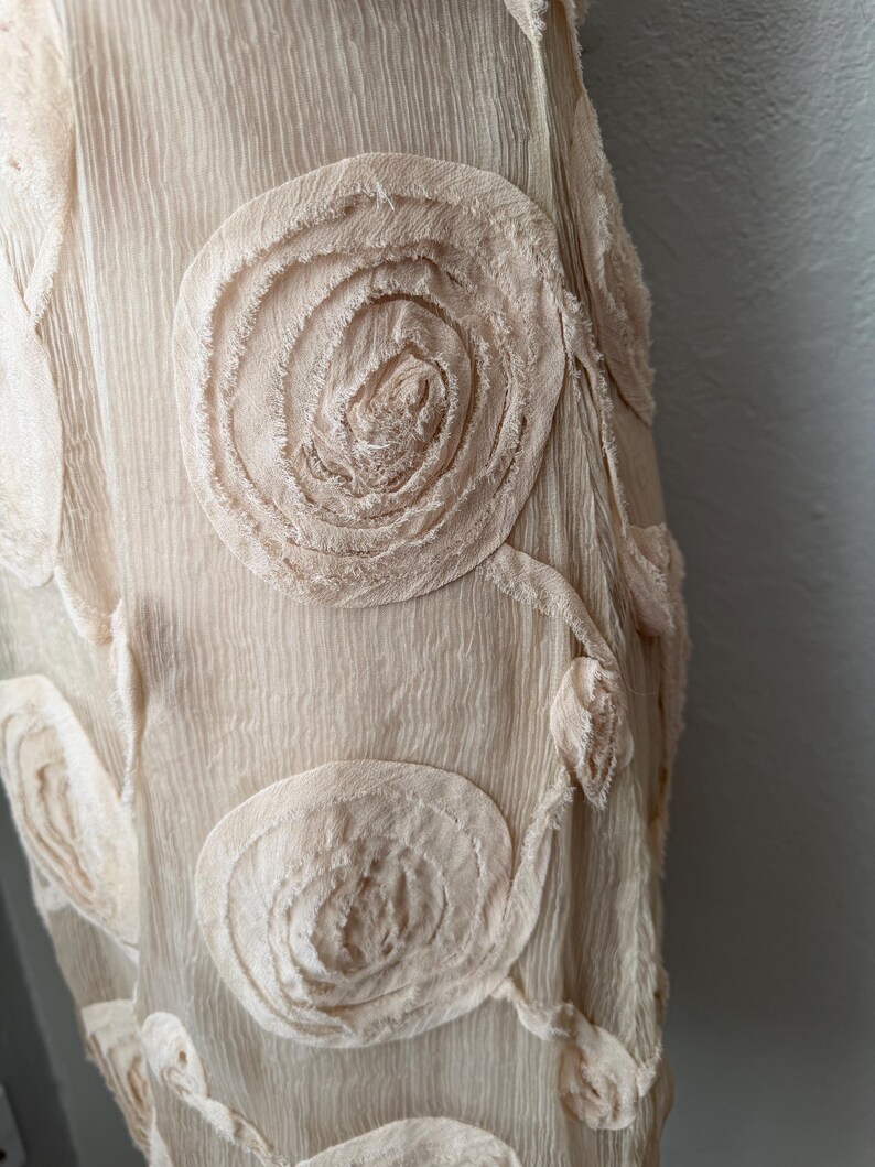 May include: Close-up of a cream-colored fabric with textured, swirling floral appliqu&eacute;s. The fabric has a crinkled appearance, and the appliqu&eacute;s are made of the same material, creating a cohesive design. The overall aesthetic is soft and delicate.