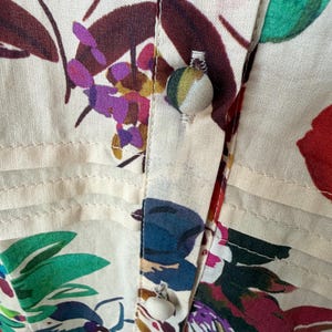 May include: Close-up of a cream-colored floral print fabric. The fabric features a vibrant pattern of red, purple, blue, and green flowers and leaves. The image highlights the buttoned placket of a shirt or blouse, showcasing the detailed stitching and fabric-covered buttons.