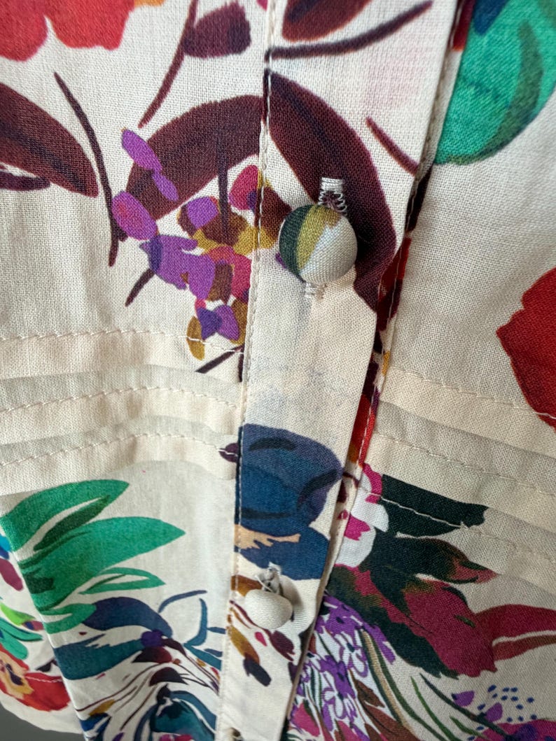 May include: Close-up of a cream-colored floral print fabric. The fabric features a vibrant pattern of red, purple, blue, and green flowers and leaves. The image highlights the buttoned placket of a shirt or blouse, showcasing the detailed stitching and fabric-covered buttons.
