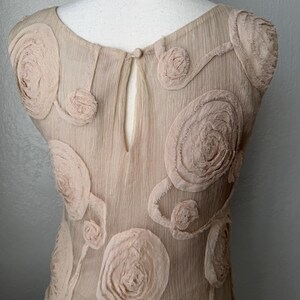 May include: A beige dress with floral embellishments. The dress features a round neckline and a keyhole back. The fabric has a textured appearance, and the floral details are in a lighter shade of beige. A gold-colored chain necklace is visible.