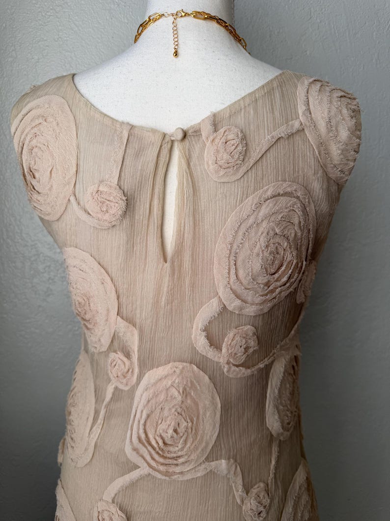 May include: A beige dress with floral embellishments. The dress features a round neckline and a keyhole back. The fabric has a textured appearance, and the floral details are in a lighter shade of beige. A gold-colored chain necklace is visible.