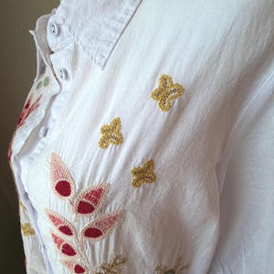 May include: White button-down shirt with embroidered floral and leaf designs in red, pink, green, and gold. The shirt features a classic collar and three gold embroidered butterfly accents.