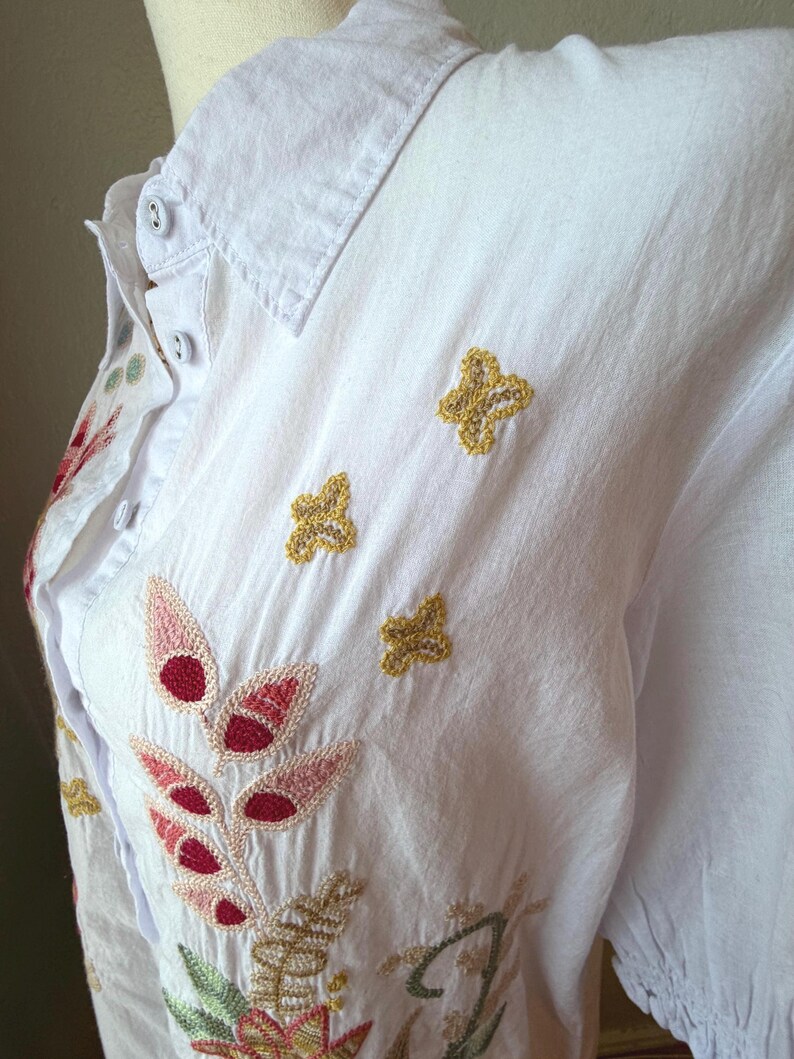 May include: White button-down shirt with embroidered floral and leaf designs in red, pink, green, and gold. The shirt features a classic collar and three gold embroidered butterfly accents.
