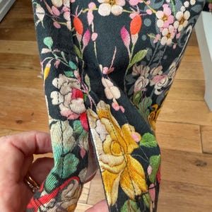 Puede incluir: Una prenda de tela negra con un estampado floral denso de flores y follaje en amarillo, rosa, blanco y verde. La tela tiene una abertura lateral visible en el dobladillo, rematada con costuras reforzadas en los bordes de la abertura.