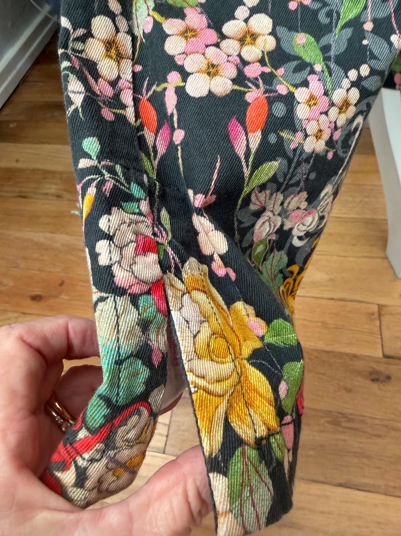 Puede incluir: Una prenda de tela negra con un estampado floral denso de flores y follaje en amarillo, rosa, blanco y verde. La tela tiene una abertura lateral visible en el dobladillo, rematada con costuras reforzadas en los bordes de la abertura.