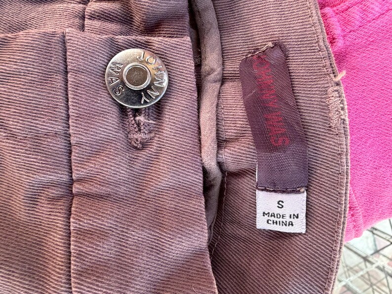 May include: Close-up of mauve fabric, possibly trousers or shorts, with a silver button reading "JOANNA WAS NY". A small purple brand tag and a white tag reading "S MADE IN CHINA" are also visible. The fabric has a textured appearance.