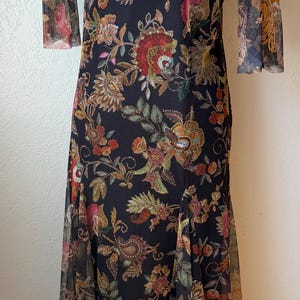 May include: A long black dress with a floral pattern. The dress has a sheer overlay with a gold and floral design on the shoulders and sleeves. The dress is displayed on a mannequin.
