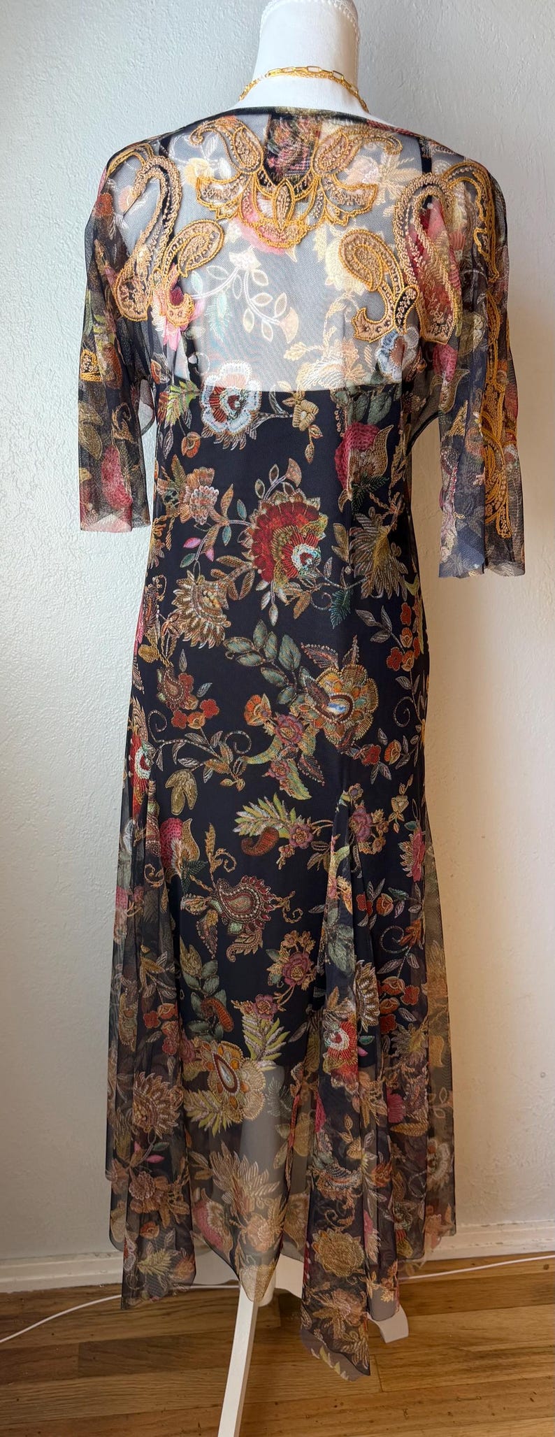 May include: A long black dress with a floral pattern. The dress has a sheer overlay with a gold and floral design on the shoulders and sleeves. The dress is displayed on a mannequin.