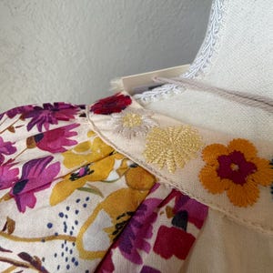 May include: Close-up of a cream-colored garment with floral print sleeves and embroidered details. The sleeves feature a vibrant pattern of purple, yellow, and pink flowers. The neckline is adorned with embroidered yellow and orange flowers.